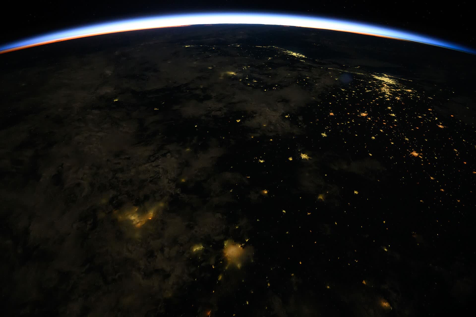 A thin blue band of sunrise along the curve of the Earth, with city lights still on below